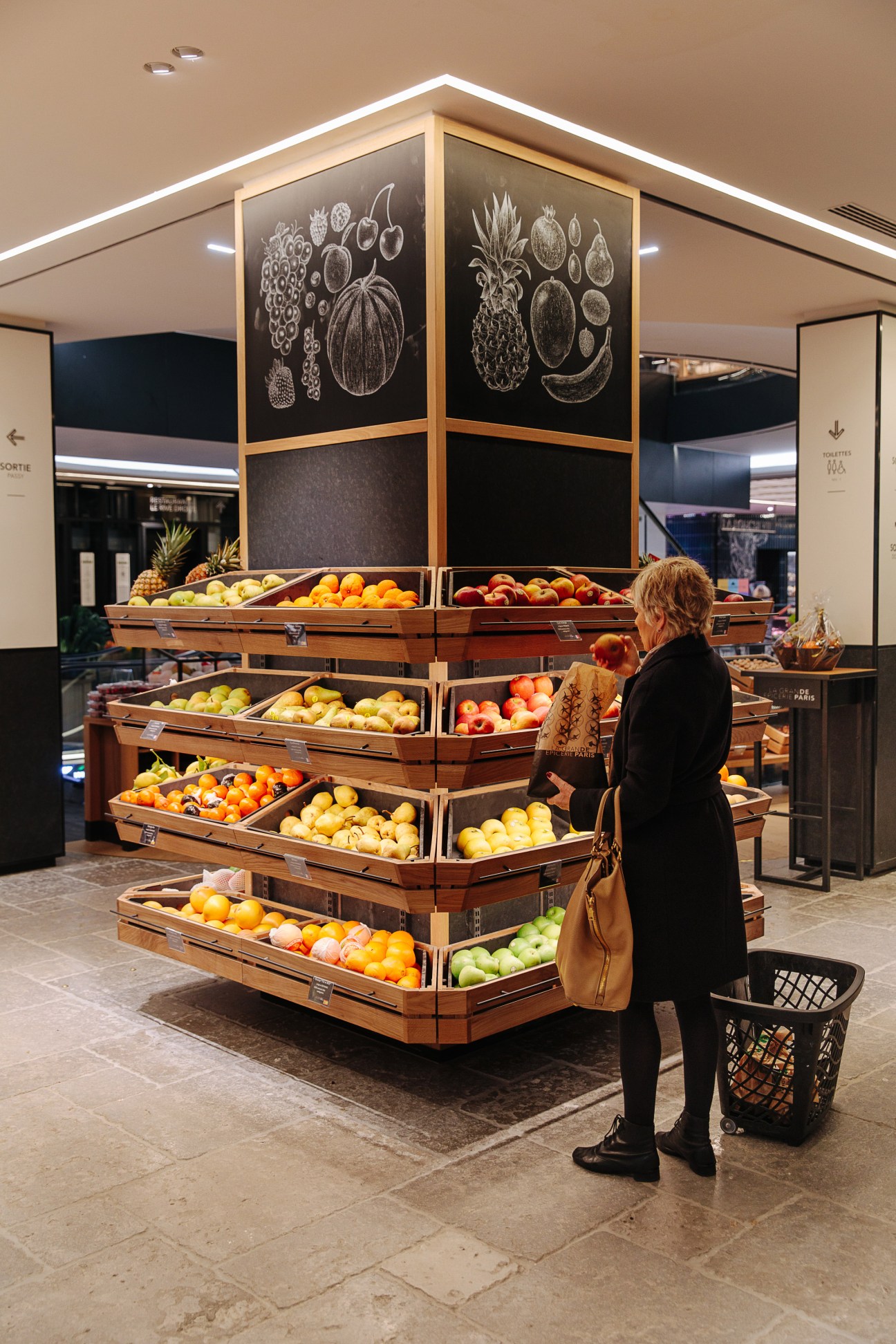 Customer browsing tiered wooden produce display with fresh fruits and illustrated fruit panels at La Grande Épicerie.