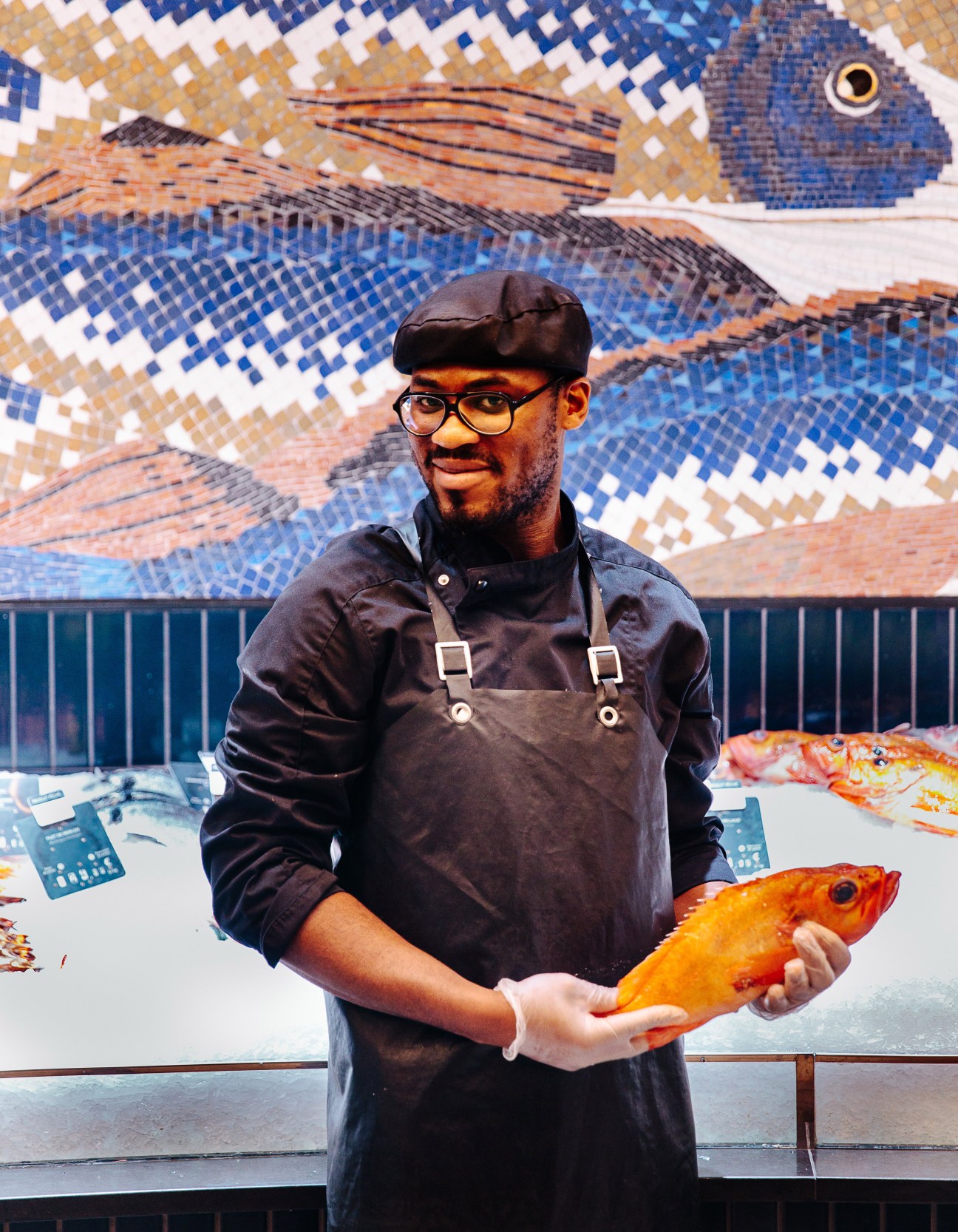 Fishmonger holding a whole fish in front of abstract glass mosaic artwork by Mathilde Jonquière.