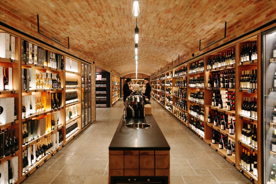 Wine cellar with wooden display cases filled with wine bottles on both sides and customers browsing in the distance.