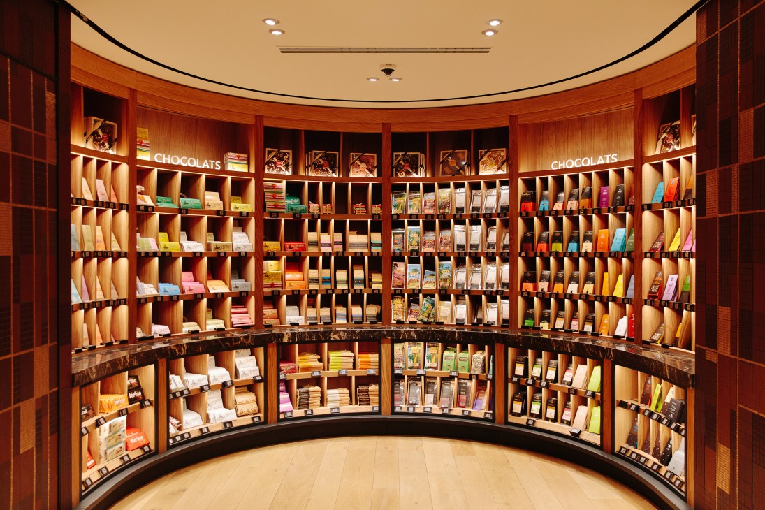 Curved wooden shelving displays various chocolate products with colorful packaging in La Grande Épicerie's chocolate emporium section.
