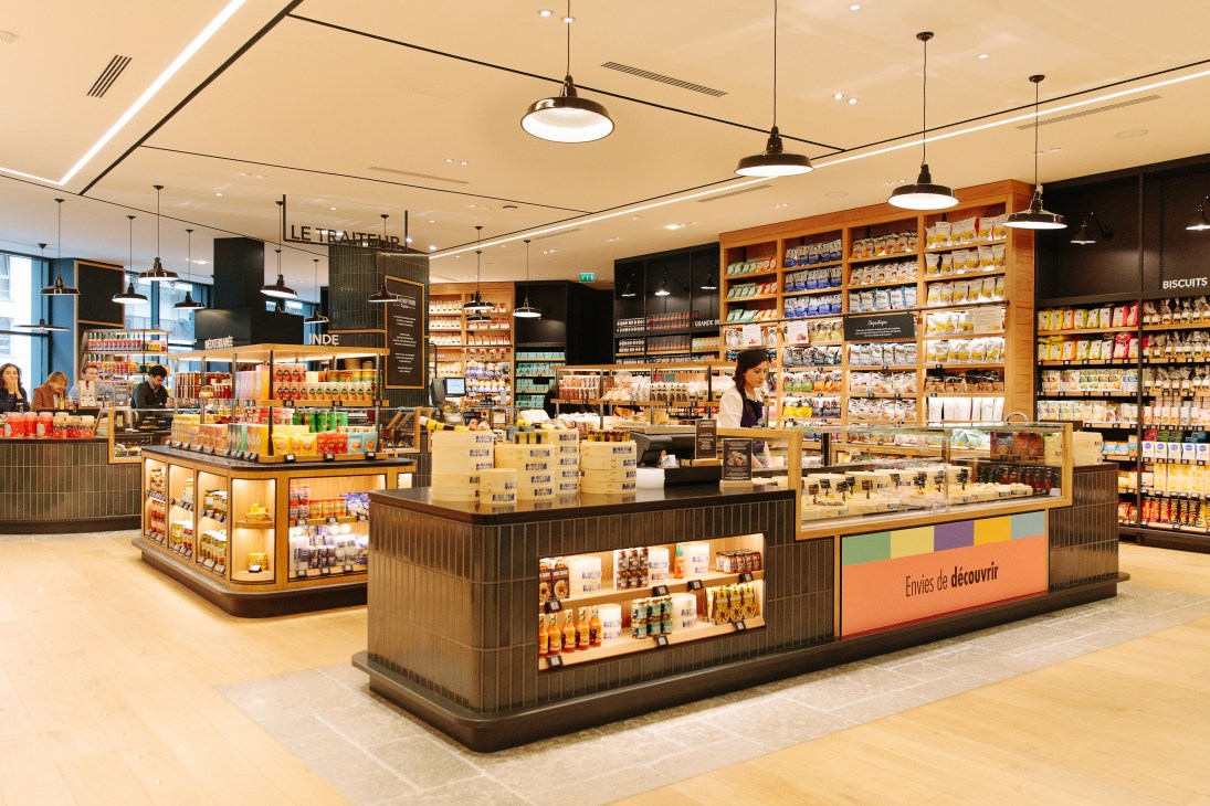 Interior of La Grande Épicerie food hall with illuminated wooden display counters, shelving stocked with packaged goods, and pendant lighting.