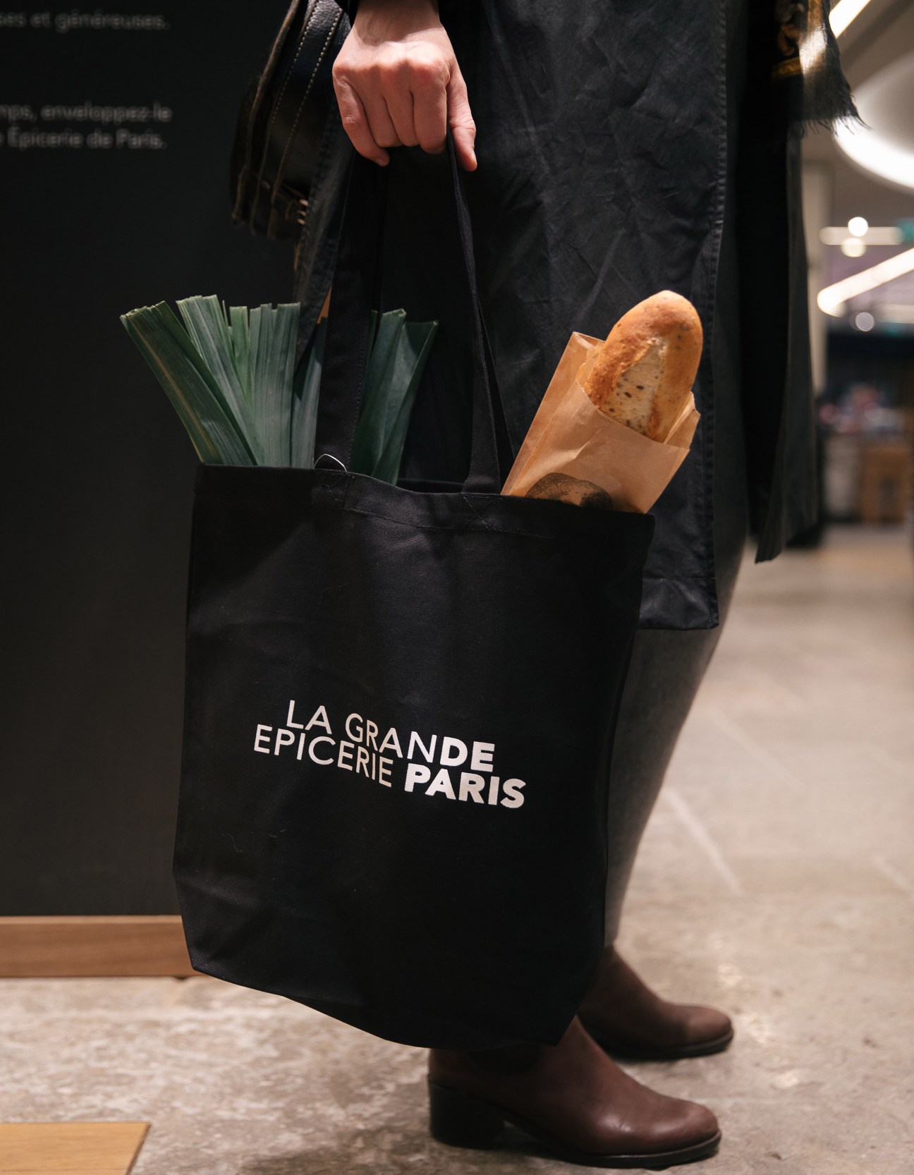 Person holding a black La Grande Épicerie Paris tote bag containing fresh leeks and a baguette.