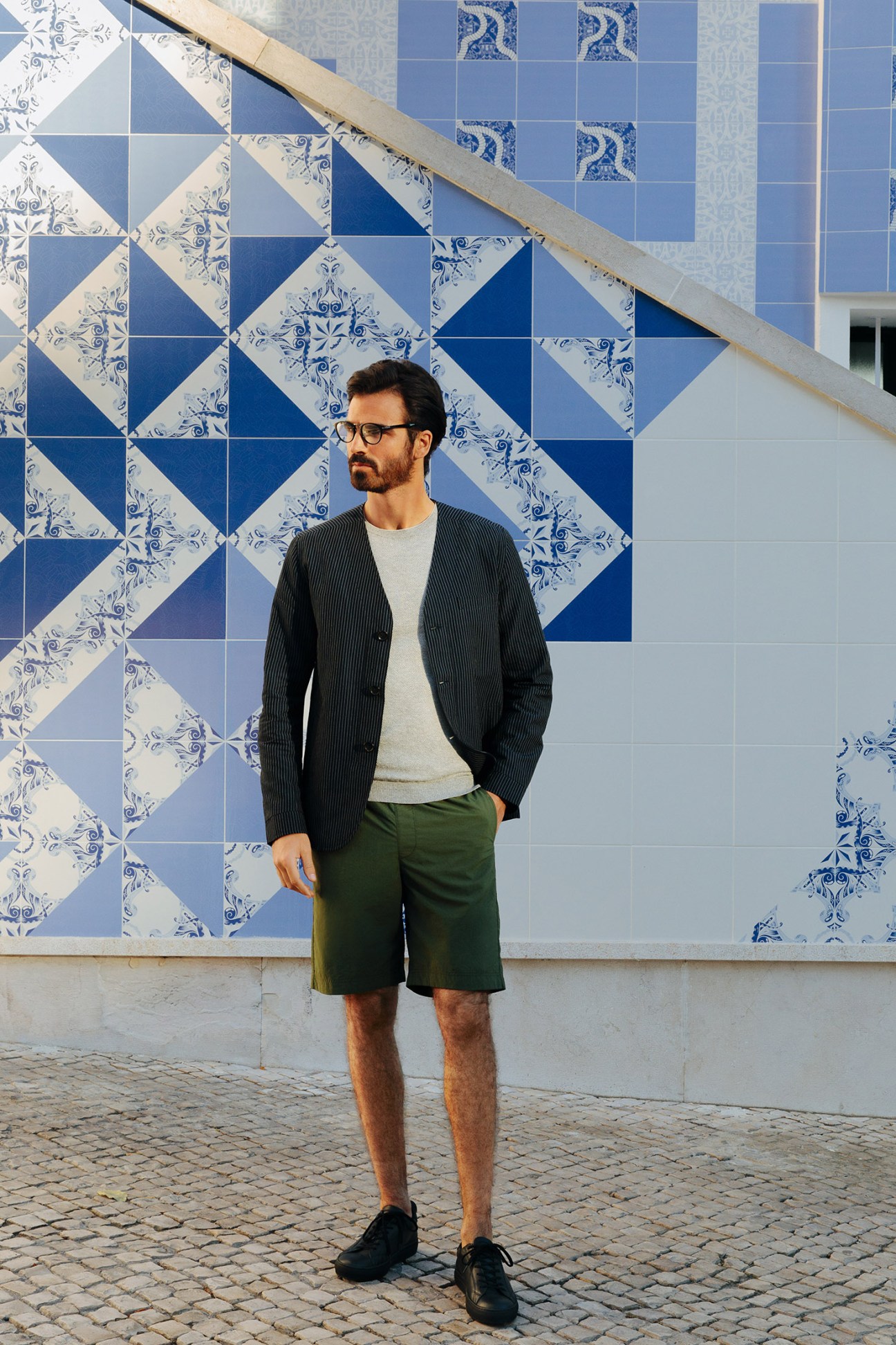 Man in Oliver Spencer jacket, Circolo 1901 jumper, and Goldwin shorts standing before blue and white patterned Portuguese tiles.
