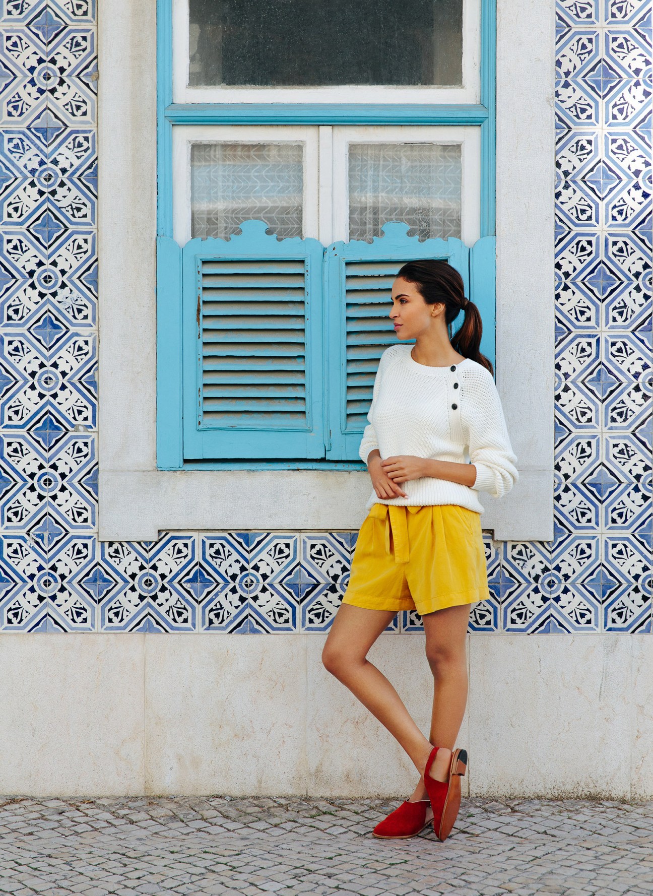 Woman in white jumper and yellow shorts standing against blue-shuttered window with patterned tiles.