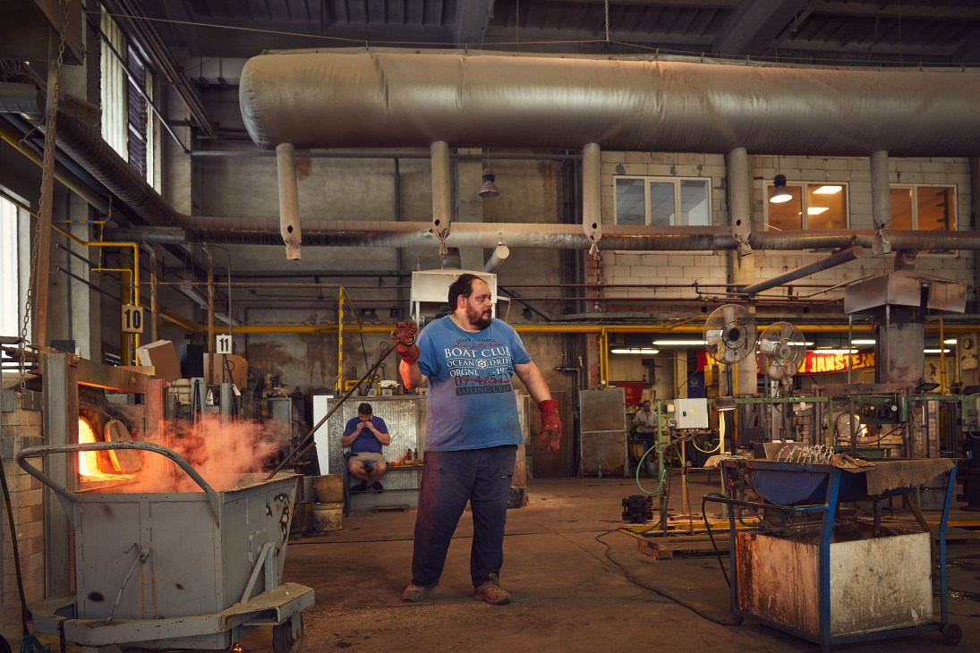 Worker in blue shirt tends to molten glass furnace in an industrial factory with overhead pipes and metal equipment.