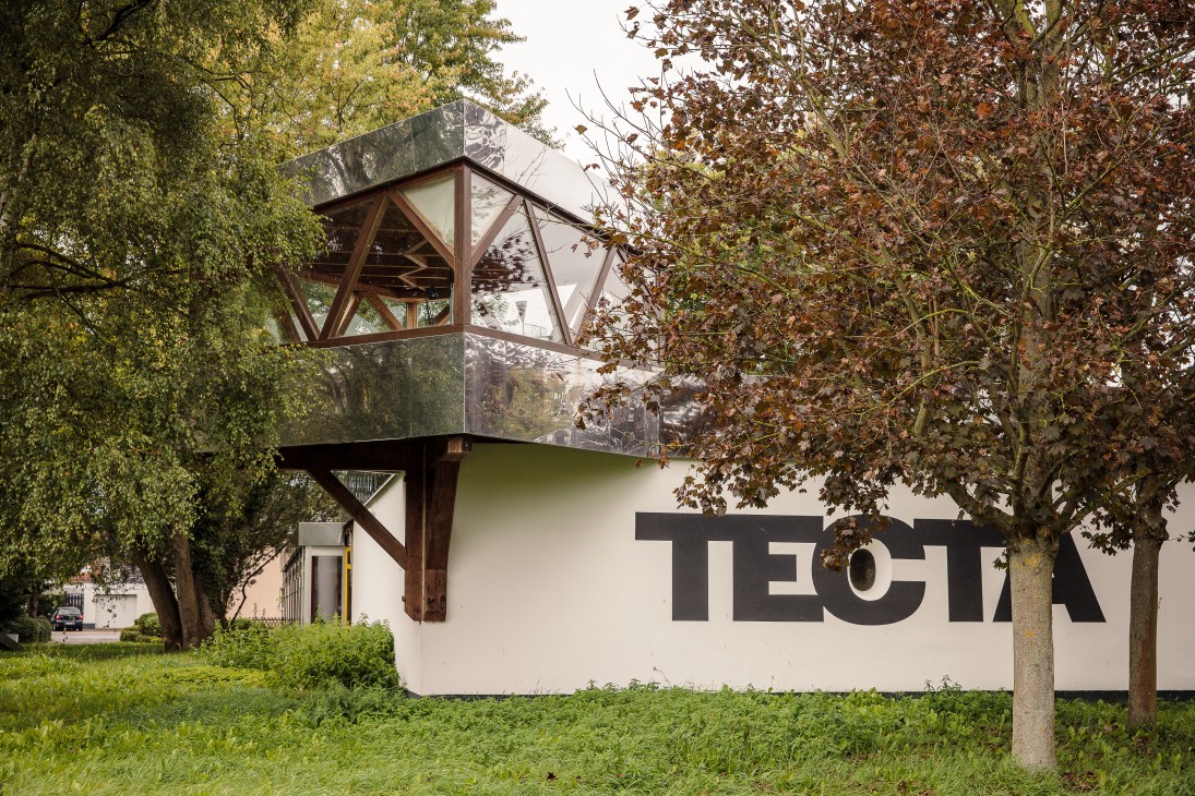 Tecta's modernist office building with white walls, geometric wooden framework, and large windows surrounded by trees.