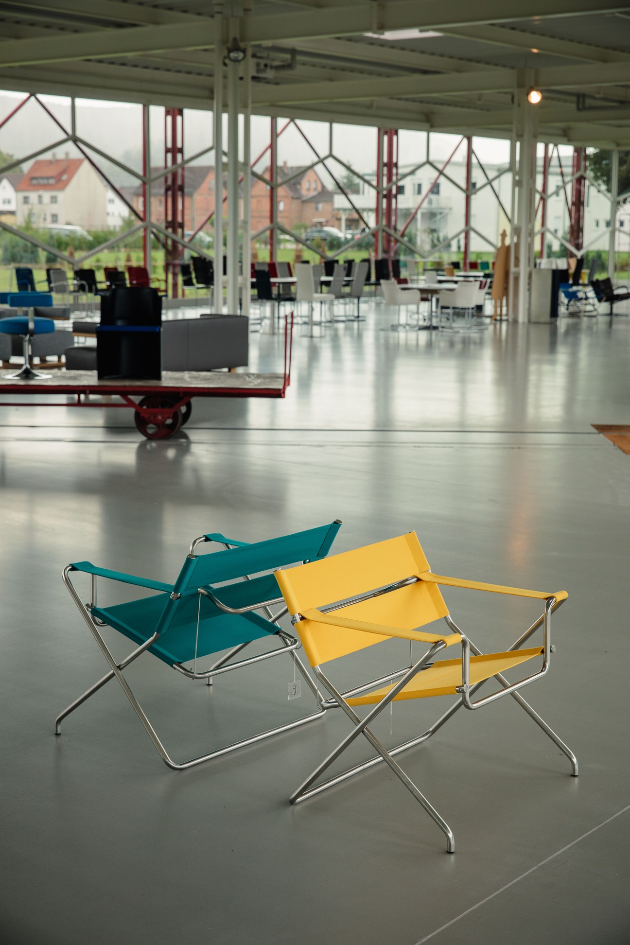 Two colorful folding camping chairs—one teal and one yellow—with metal frames on a polished concrete floor in a bright modernist interior space.