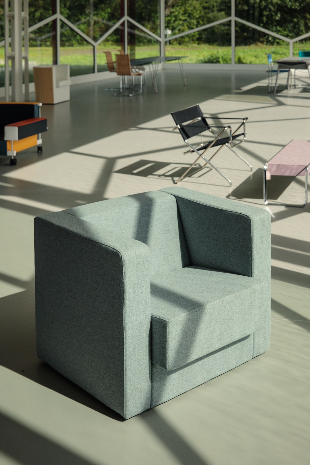 A light green upholstered modernist armchair displayed in a sunlit gallery space with geometric lattice windows and various chairs visible in the background.