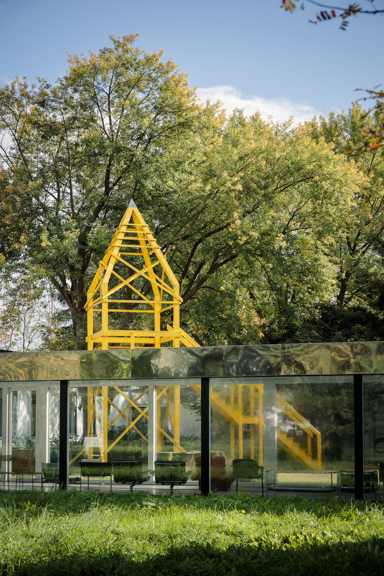 Yellow geometric lattice structure on modernist building with glass windows and autumn trees behind.