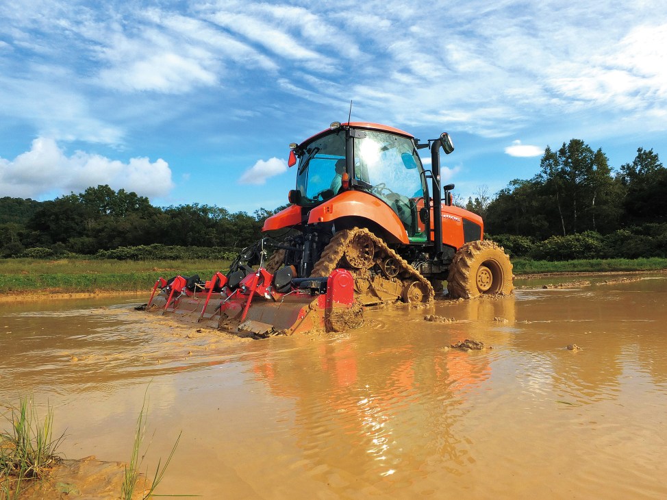 Orange Kubota MR97 tractor with red farming implements working in muddy water during field operations.