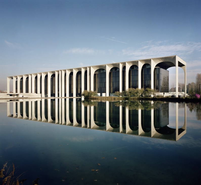 Palazzo Mondadori's modernist concrete façade with distinctive arches reflects in a still lake beneath clear sky.