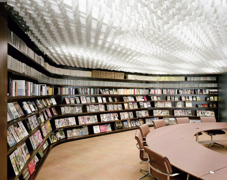 Modern boardroom with curved bookshelves, geometric ceiling grid, and wooden table with tan chairs.