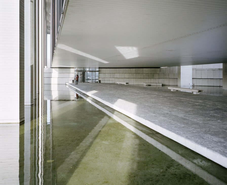 A minimalist elevated concrete walkway with railings beneath a white ceiling, overlooking a reflecting pool with natural light streaming in.