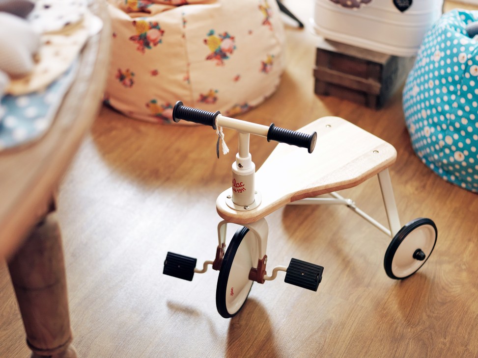A wooden children's tricycle with black handlebars and pedals on a wooden floor with toys in the background.