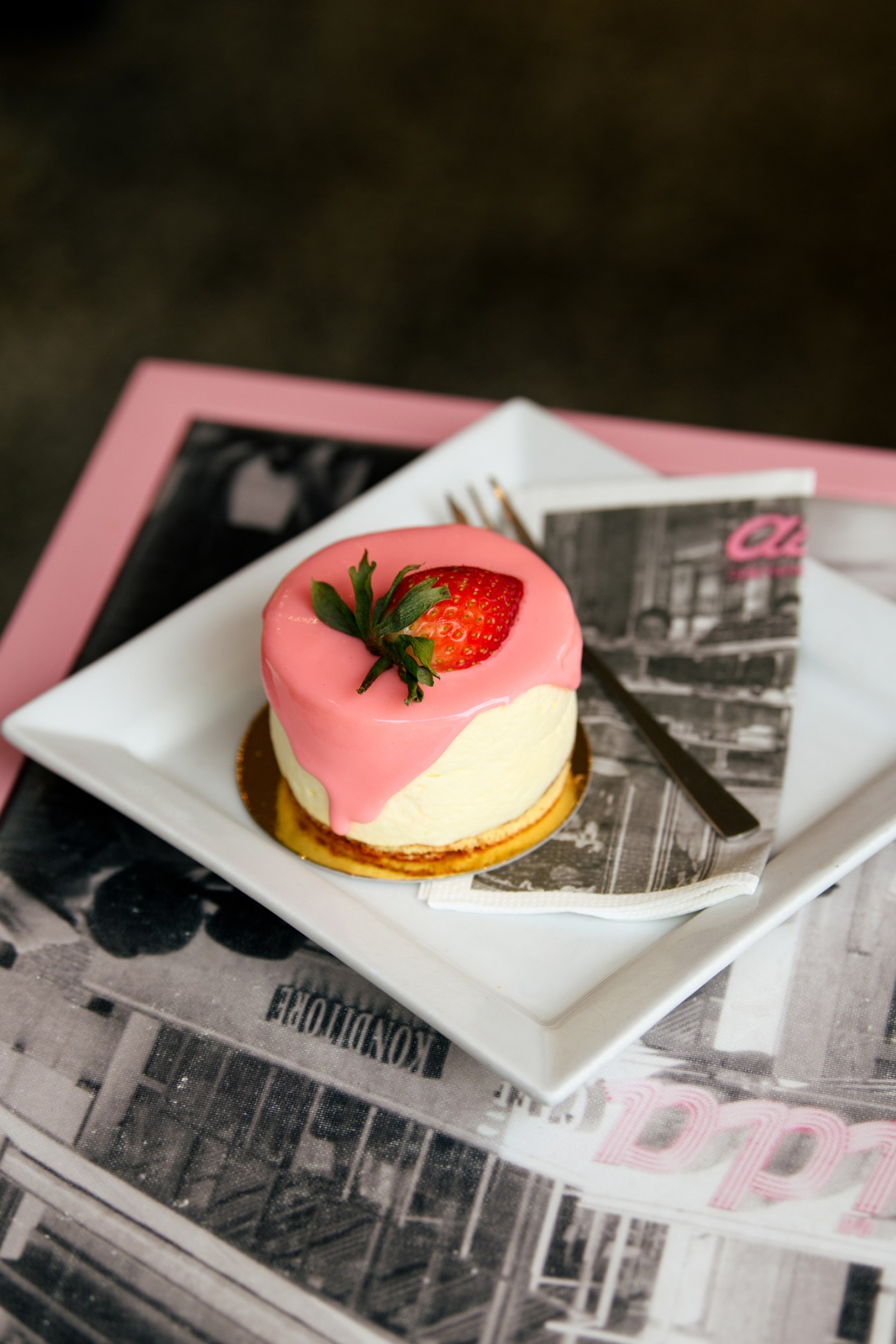 Pink and white layered cheesecake tartlet topped with fresh strawberry on white square plate with fork and magazines