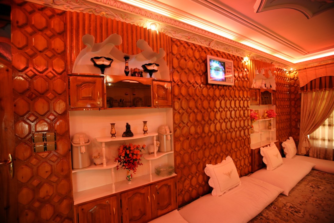 An ornate sitting room with red-orange wood panelling, geometric wall patterns, shelving units with decorative items, and red ambient lighting throughout.