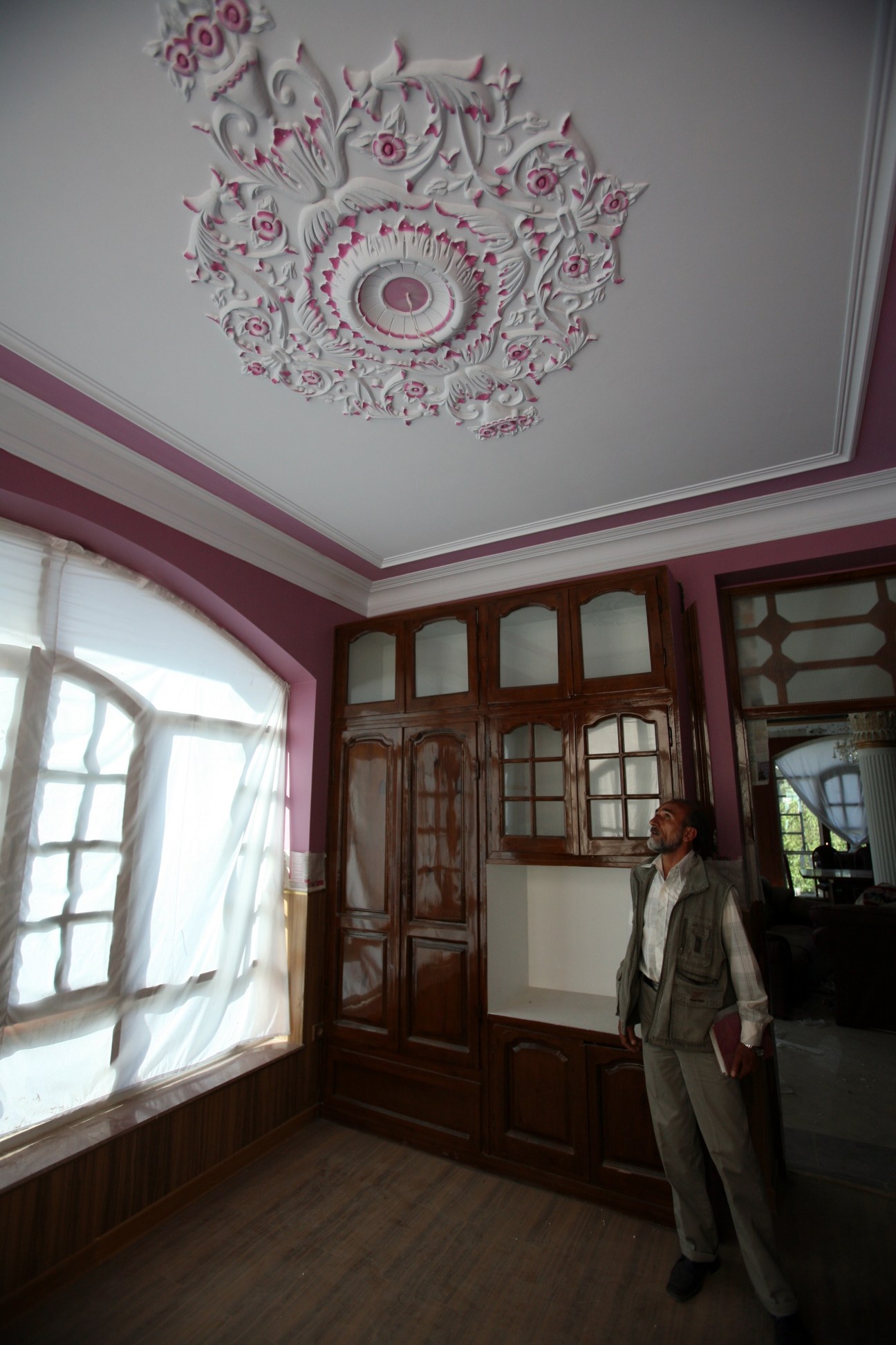 Man in light jacket stands in ornate room with pink and white decorative ceiling rosette and wooden doors.