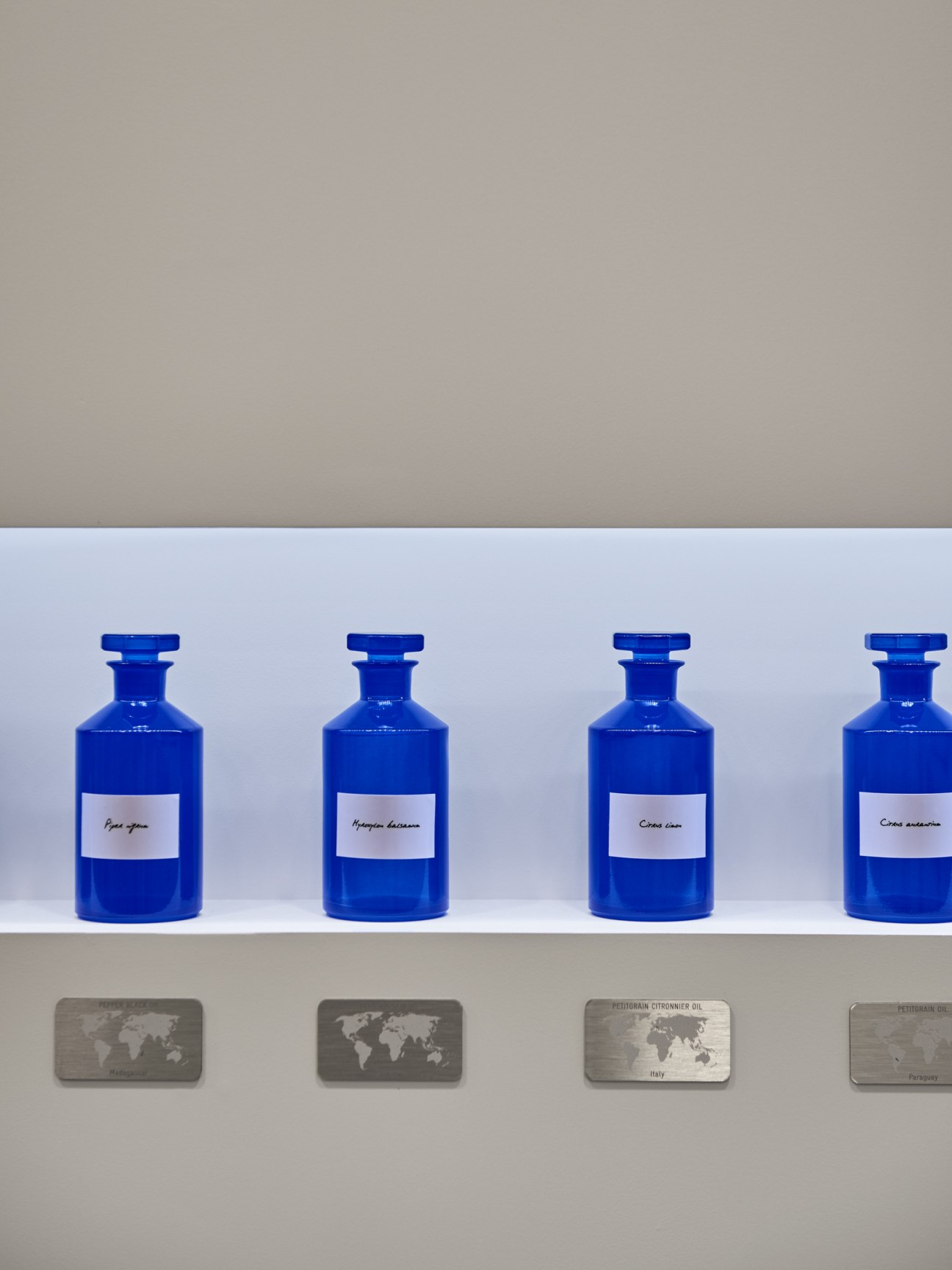 Four blue glass vials with white labels containing fragrance ingredients, displayed on a shelf with geographic origin markers below.
