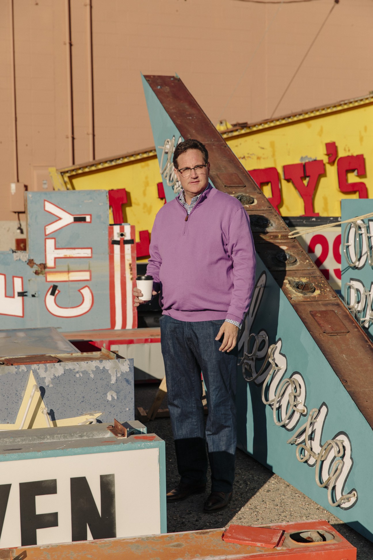 Ed Garcia stands among vintage neon signs from Route 66, wearing a purple pullover and holding a coffee cup.