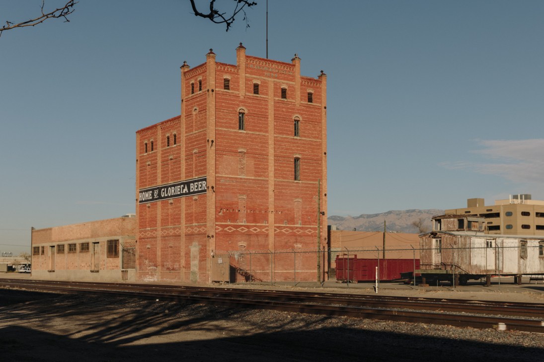 Red brick historic tower building with 'Home of Glorieta Beer' sign, railroad tracks in foreground, Albuquerque mountains visible in background.