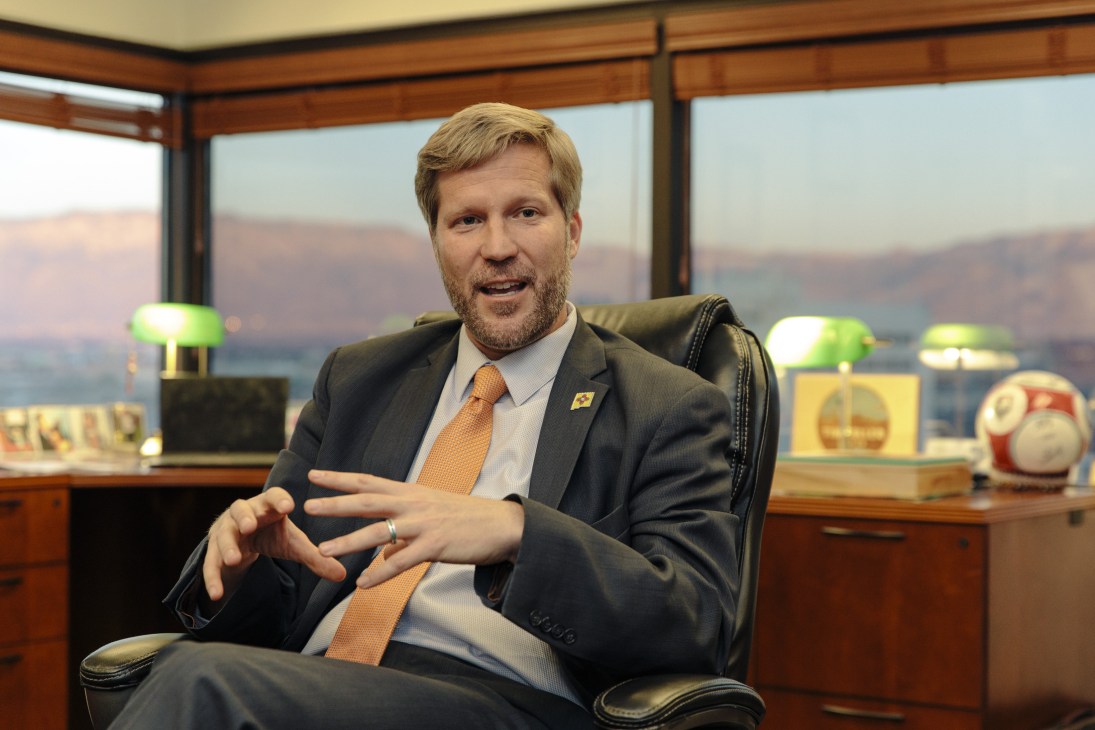 Mayor Tim Keller sitting in his downtown office with the Sandia Mountains visible through the windows behind him.