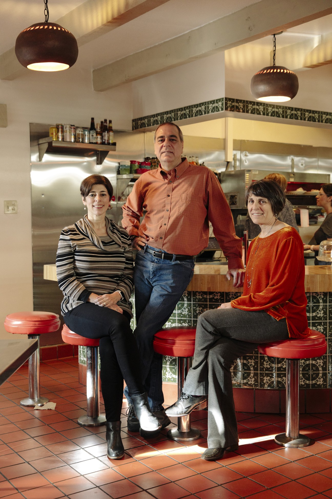 Three restaurateurs standing together in a modern kitchen with red bar stools and decorative tile backsplash.