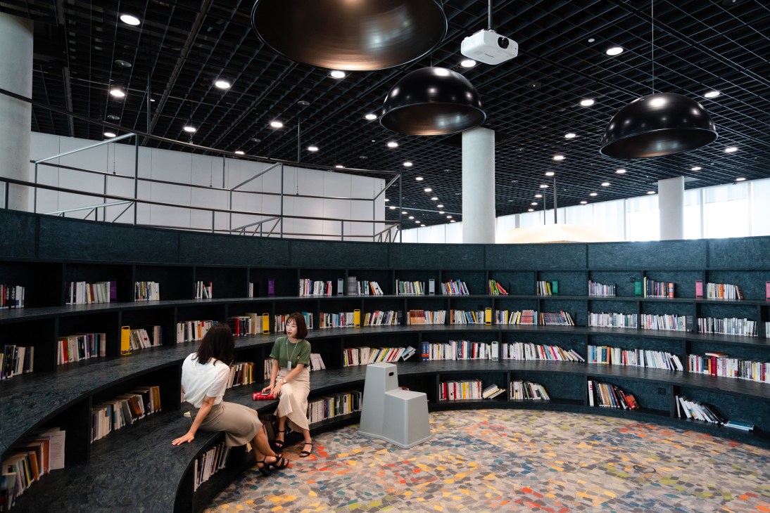Two people sit in a curved library with dark shelving filled with books, beneath black pendant lamps in a modern office space.