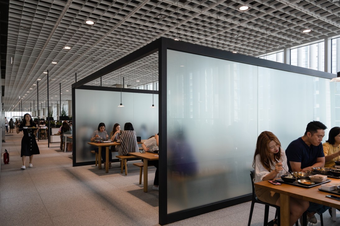 Amorepacific employees dining in an open, modern cafeteria with black-framed glass partitions and wooden tables.