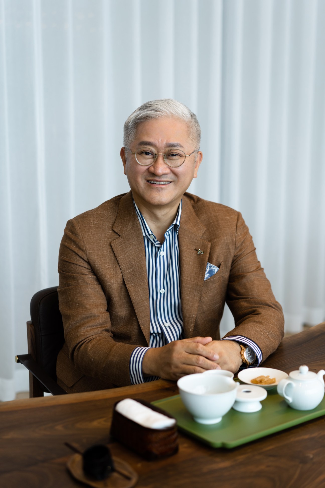 Amorepacific CEO Suh Kyung-bae sitting at a table with tea service, wearing a brown blazer and striped shirt.