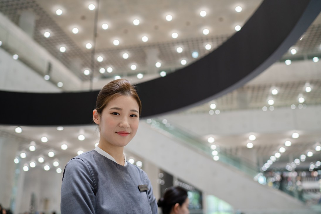 Woman in blue uniform stands in modern lobby with curved ceiling and recessed lighting at Amorepacific headquarters.