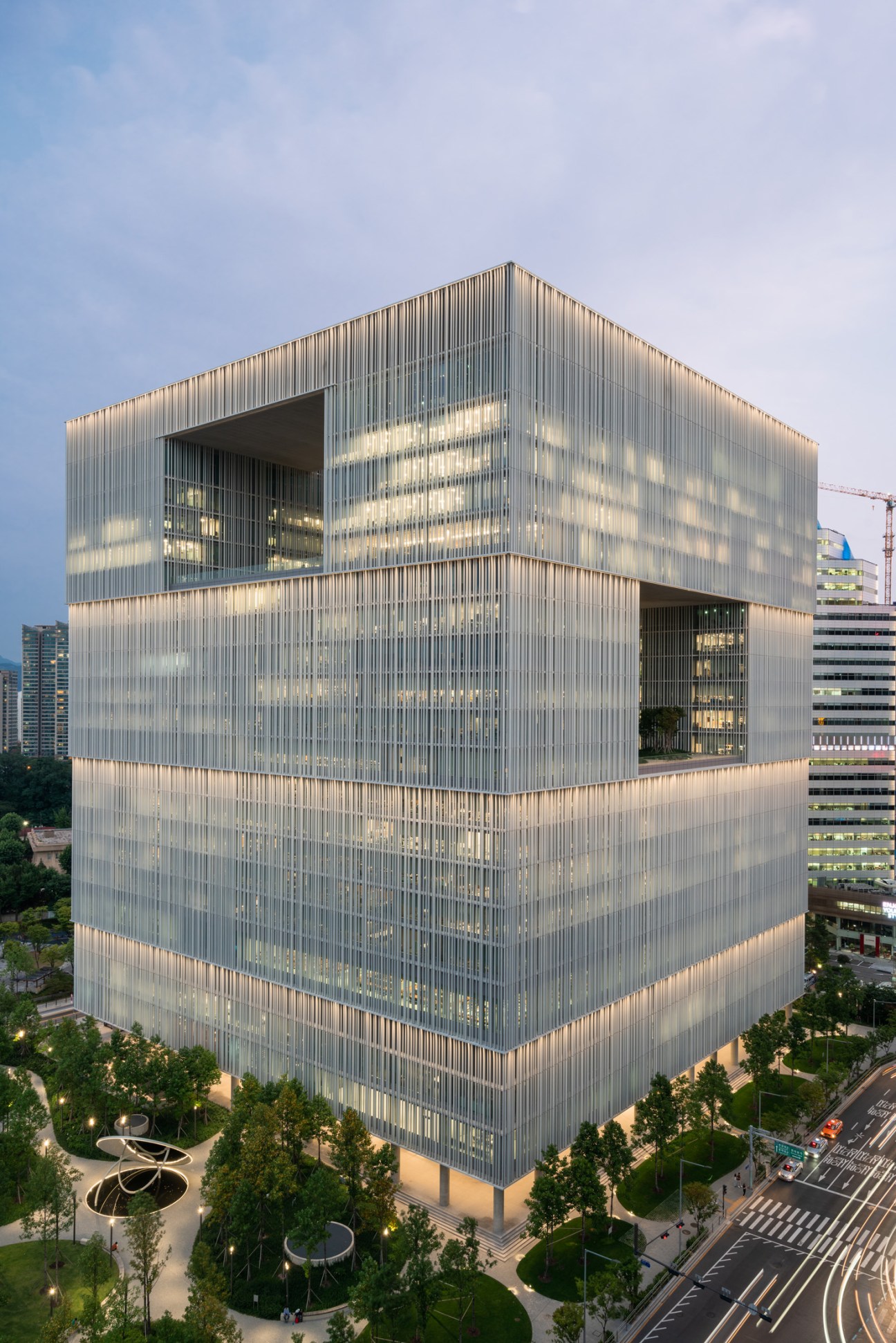 Amorepacific's 23-storey cubic headquarters building with vertical metal screening and illuminated interior, surrounded by trees and Seoul cityscape.