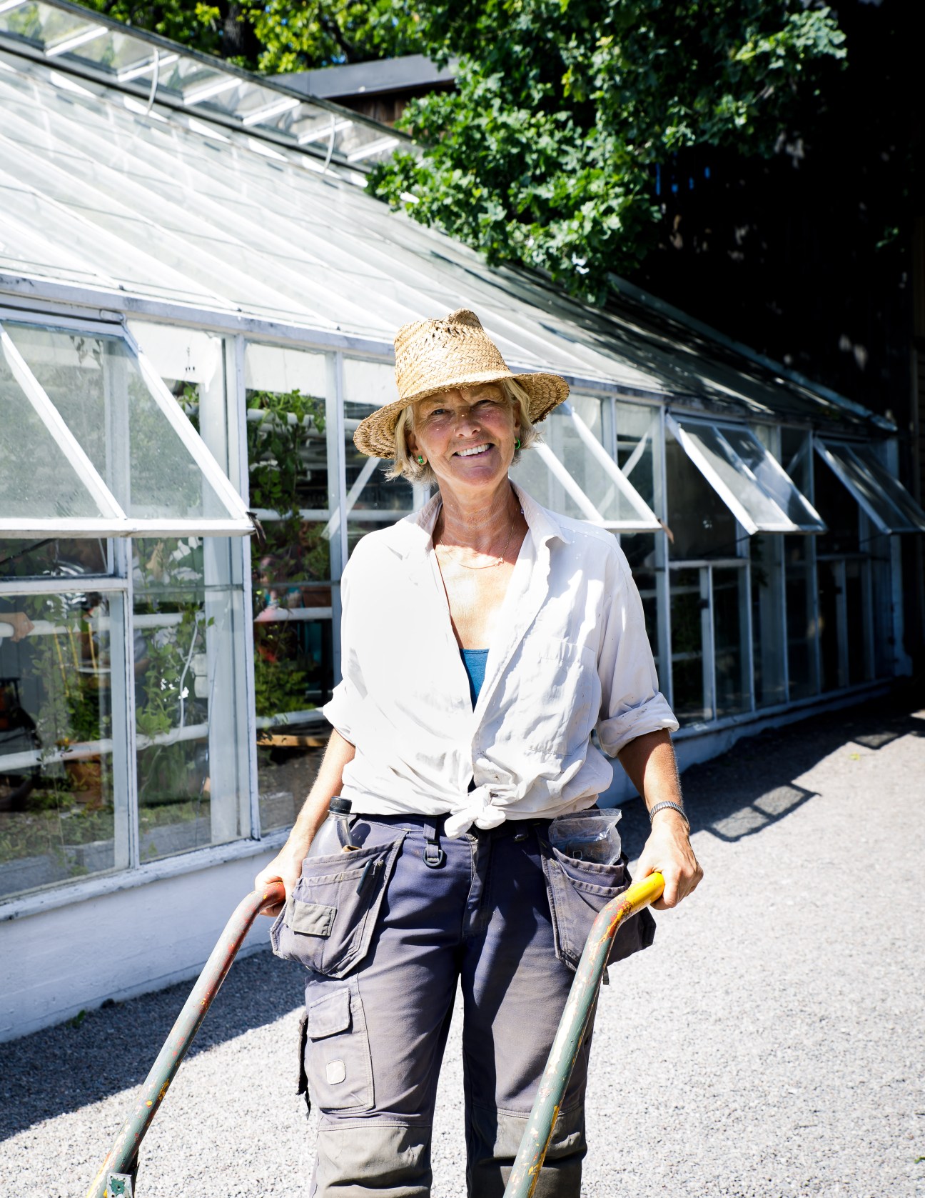 Woman in straw hat and white shirt standing with wheelbarrow handles outside greenhouse