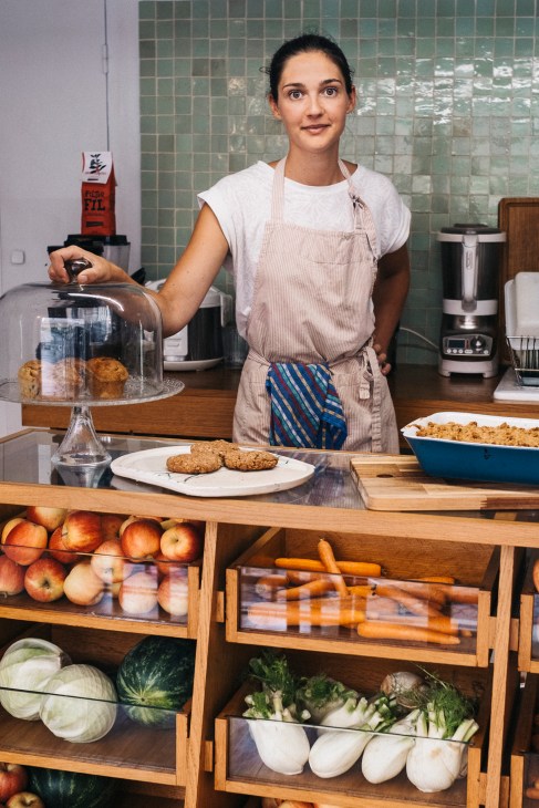 Woman in apron standing behind counter at La Piscine Kamouraska with fresh produce and baked goods displayed.