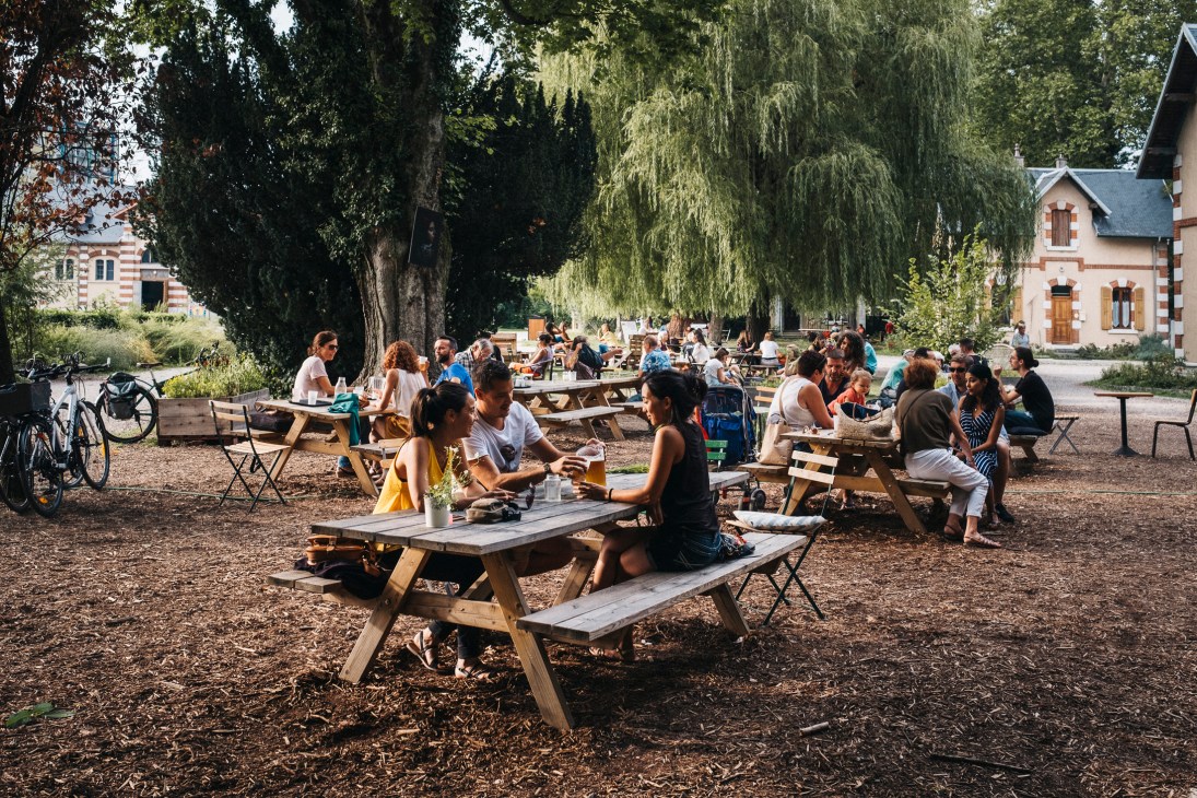 People gathering at outdoor picnic tables under large willow trees at the Haras in Annecy.