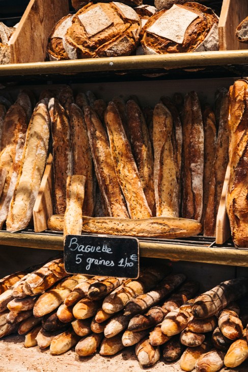 Artisanal baguettes and bread loaves displayed in wooden shelves at a French bakery with a price sign reading 'Baguette 5 graines 1/10'.