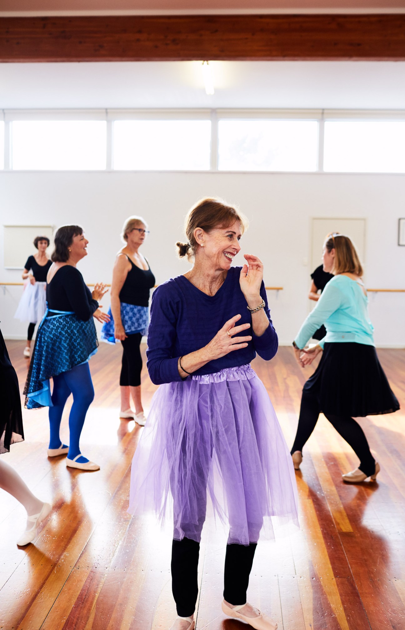 Senior ballet dancers in a studio class, with a woman in a purple tutu smiling in the foreground.