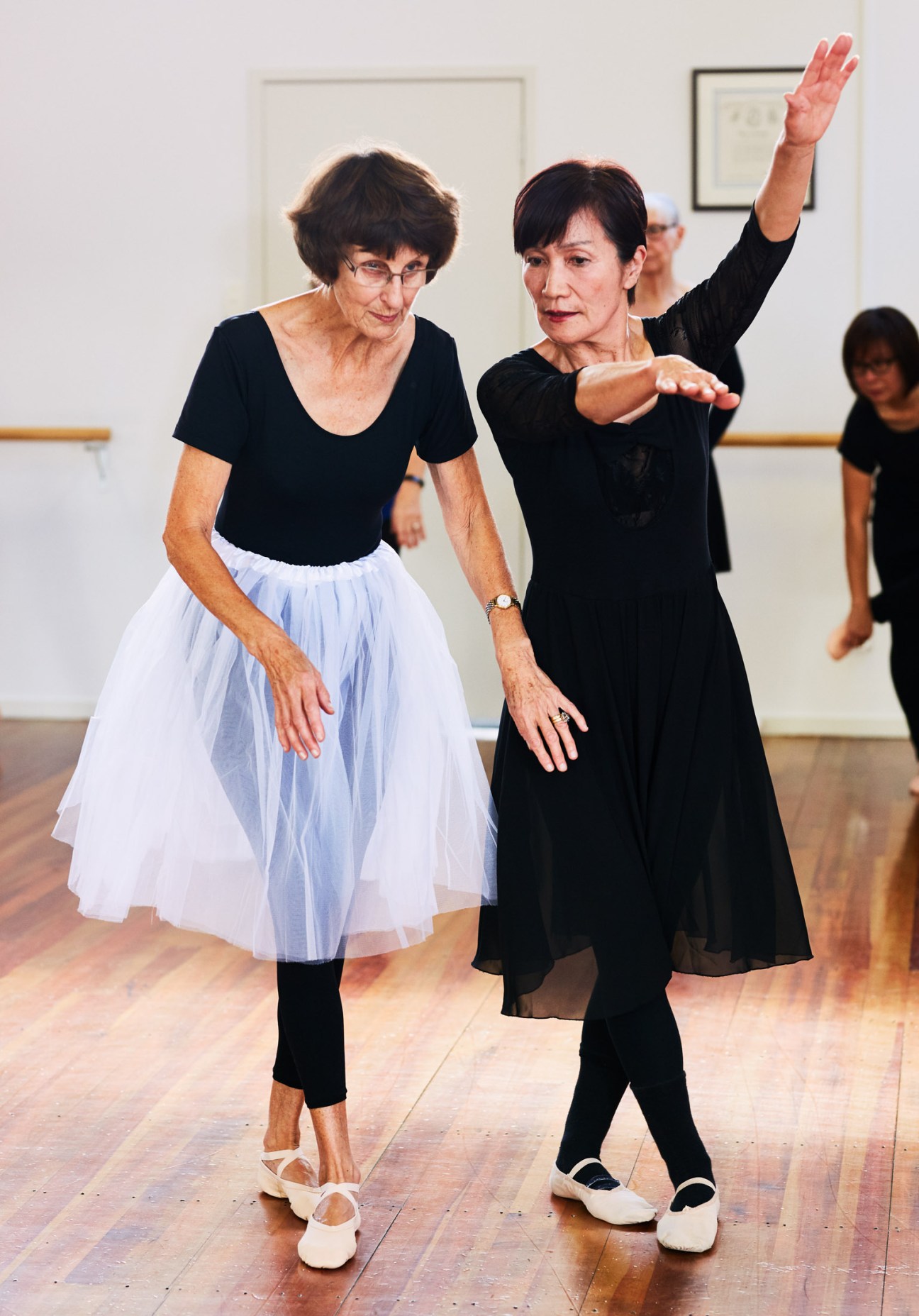 A ballet instructor demonstrates a movement to a senior student during a dance class at the barre.