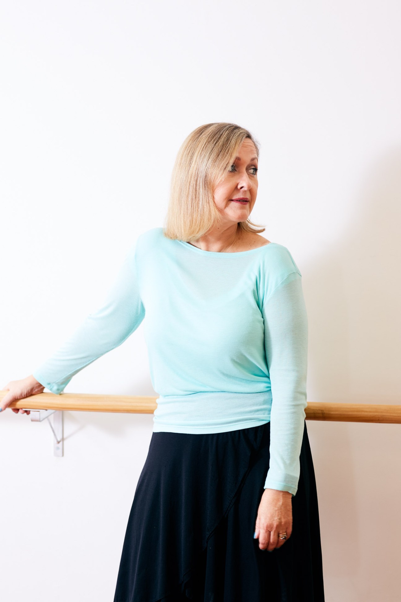 Kathleen Curwen-Walker rests her hand on the ballet barre while wearing a light blue long-sleeved top and dark skirt.