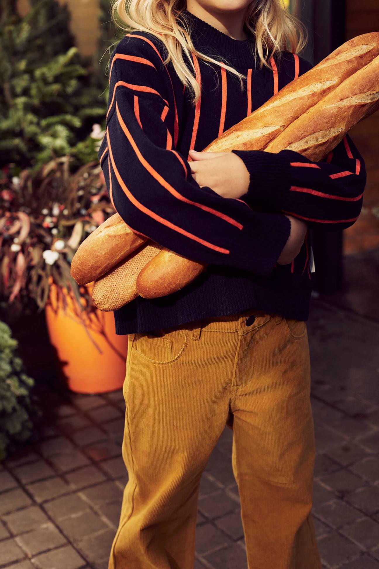 Child wearing navy striped sweater and mustard corduroy pants holding fresh baguettes