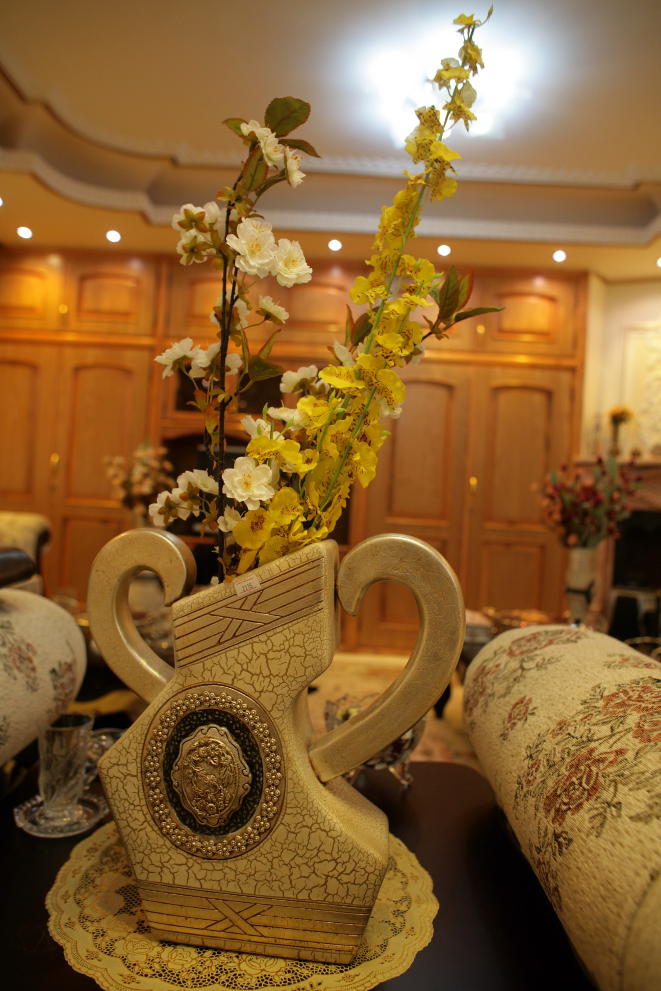 Artificial white and yellow flowers arranged in an ornate cream-colored vase with handles and decorative medallion detail.