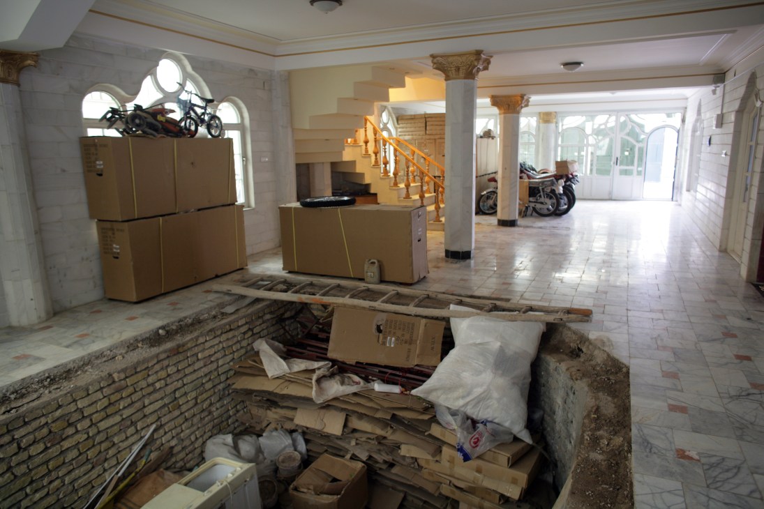 A large unfinished basement space with exposed brick, construction materials, cardboard boxes, motorcycles, and ornate columns under renovation.