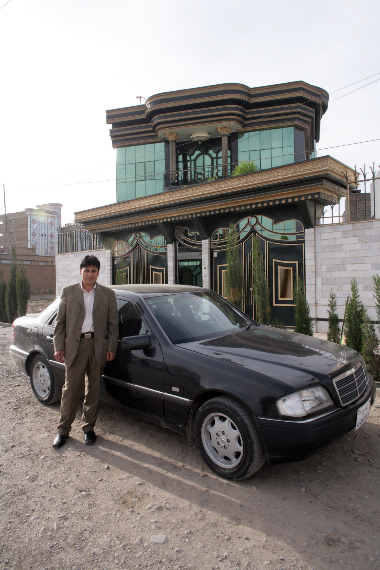 Mohammad Zaher Faqiry stands beside a black Mercedes outside his ornate three-storey mansion in Herat with mirrored glass facade and decorative architectural details.