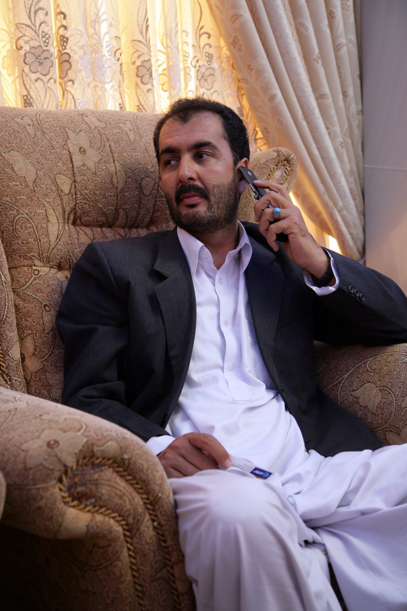 Man in dark blazer and white shirt sits on ornate beige sofa talking on phone with patterned curtains behind.