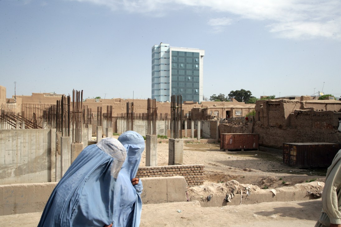 Woman in blue burqa walks past modern office building under construction in Herat.