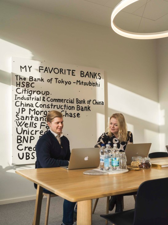 Two people work at a wooden table in a bright office with a pop art painting listing banks on the white wall behind them.