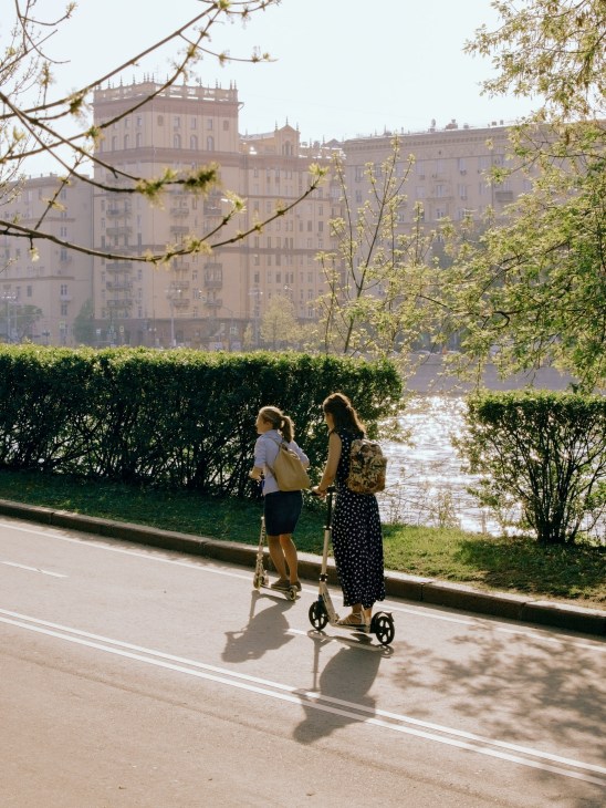 Two young women riding scooters along a tree-lined riverside path with Soviet-era apartment buildings in the background.