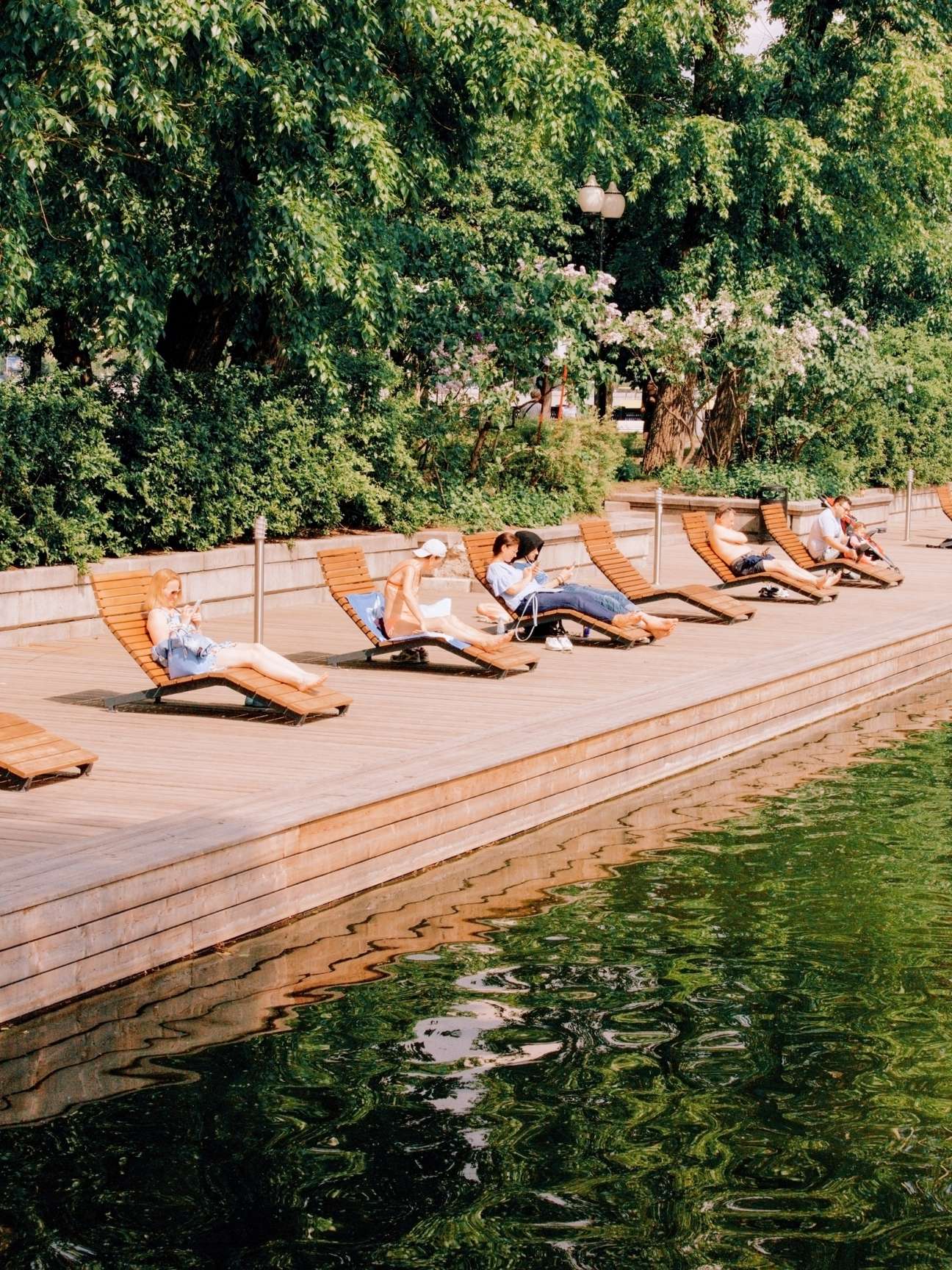 People relaxing on wooden chaise longues beside a pond in Neskuchny garden, Moscow.