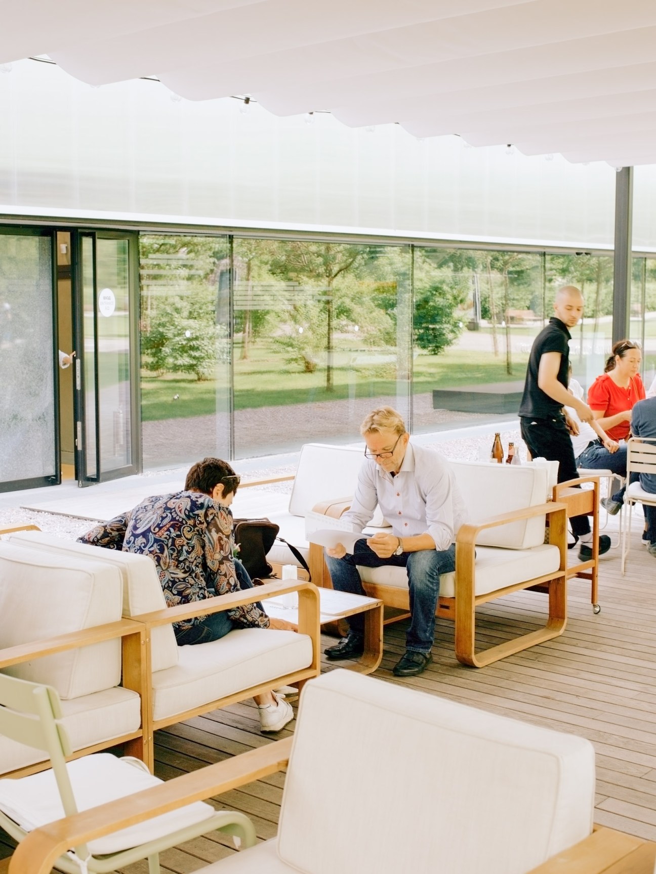 People relaxing on wooden-framed chairs with white cushions on a modern café terrace with glass walls overlooking green grounds.