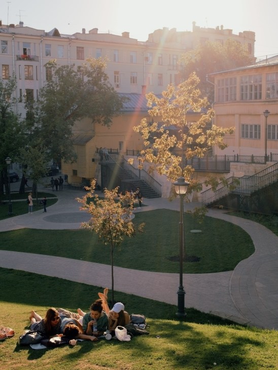 People relaxing on grass in a renovated Moscow square with historic buildings and autumn trees.