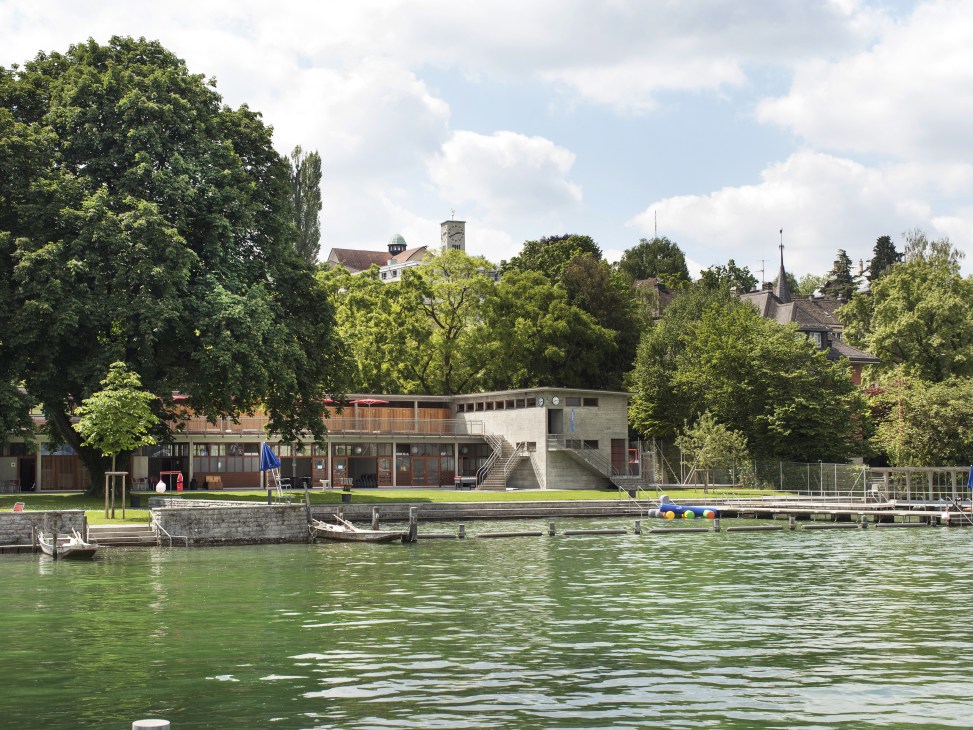 Strandbad Wollishofen swimming complex on Lake Zürich with modernist building, green lawn, and Zürich cityscape beyond.